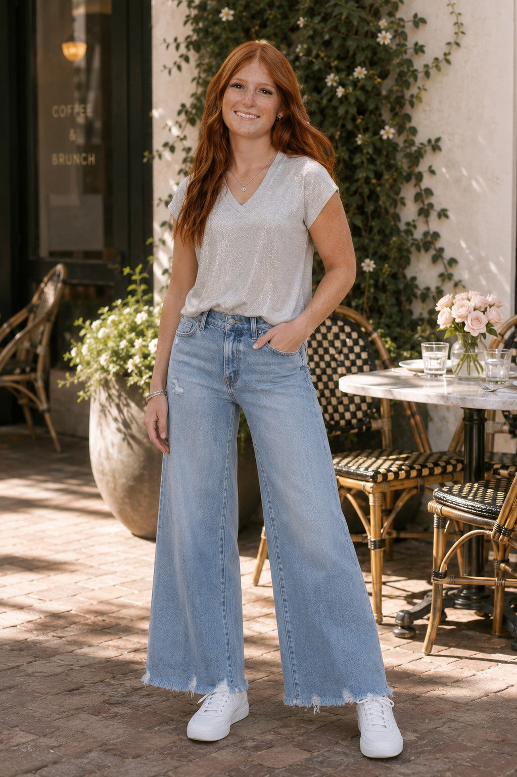 Redhead model wearing a silver metallic shimmer V-neck top with light wash distressed wide leg jeans, silver jewelry, and white sneakers at a sunny outdoor brunch café.