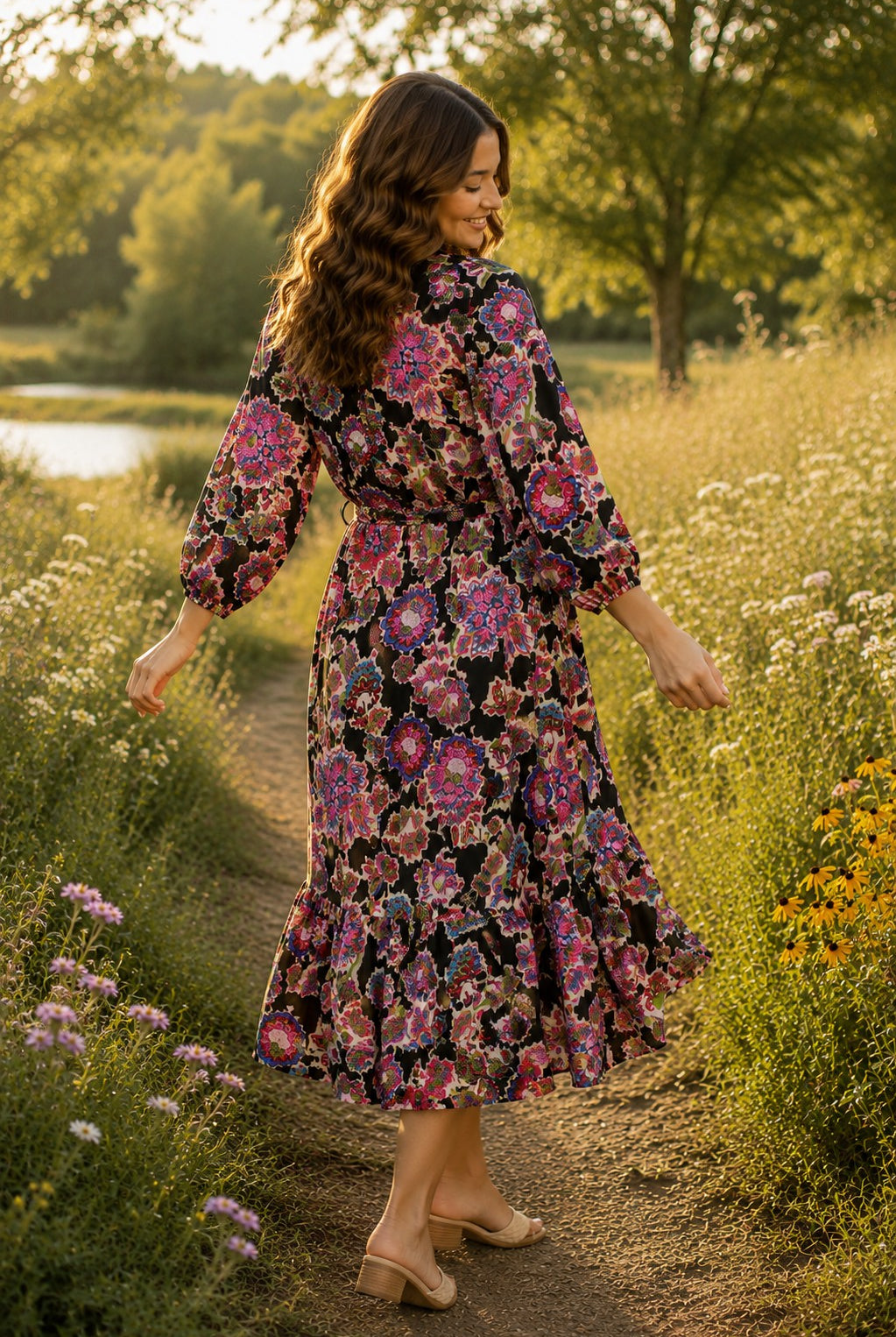 Back view of a brunette model wearing the Umgee Exclusive black floral midi dress with a belted waist, flowing midi skirt, and vibrant embroidered-inspired floral print, walking through a golden late summer meadow surrounded by wildflowers and soft evening sunlight in a carefree lifestyle setting.