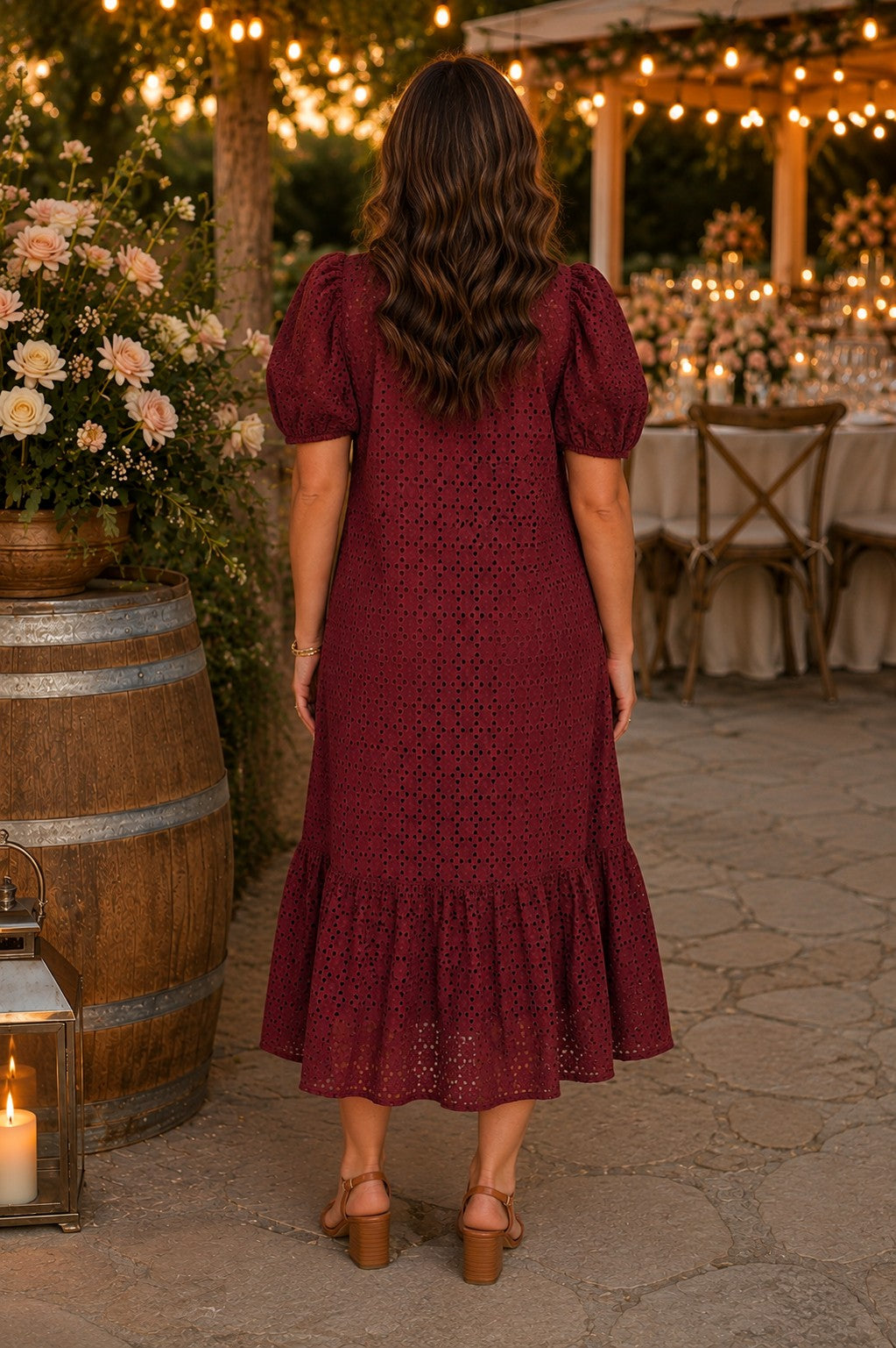 Back view of a model wearing the Entro burgundy eyelet puff sleeve midi dress with tan block heel sandals at a romantic late summer outdoor wedding venue.