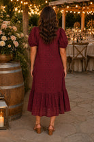 Back view of a model wearing the Entro burgundy eyelet puff sleeve midi dress with tan block heel sandals at a romantic late summer outdoor wedding venue.