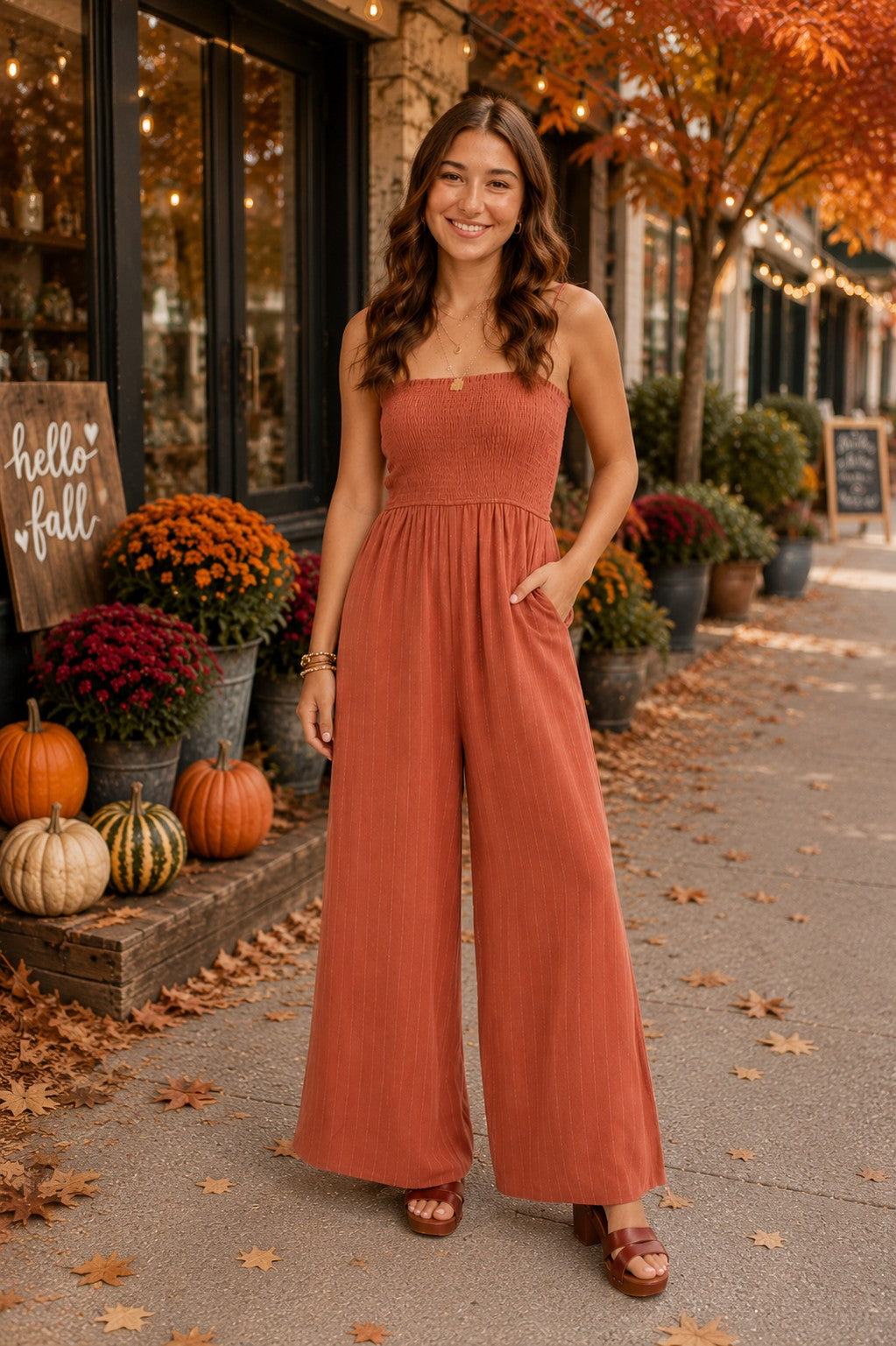 Laney modeling a mahogany smocked wide-leg jumpsuit with subtle pinstripes in a charming fall downtown setting featuring pumpkins, mums, autumn leaves, and warm seasonal décor.