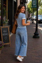 Back and side view of model wearing a blue and white striped scalloped short sleeve knit top with light wash cropped wide leg jeans while walking through a charming downtown shopping district.